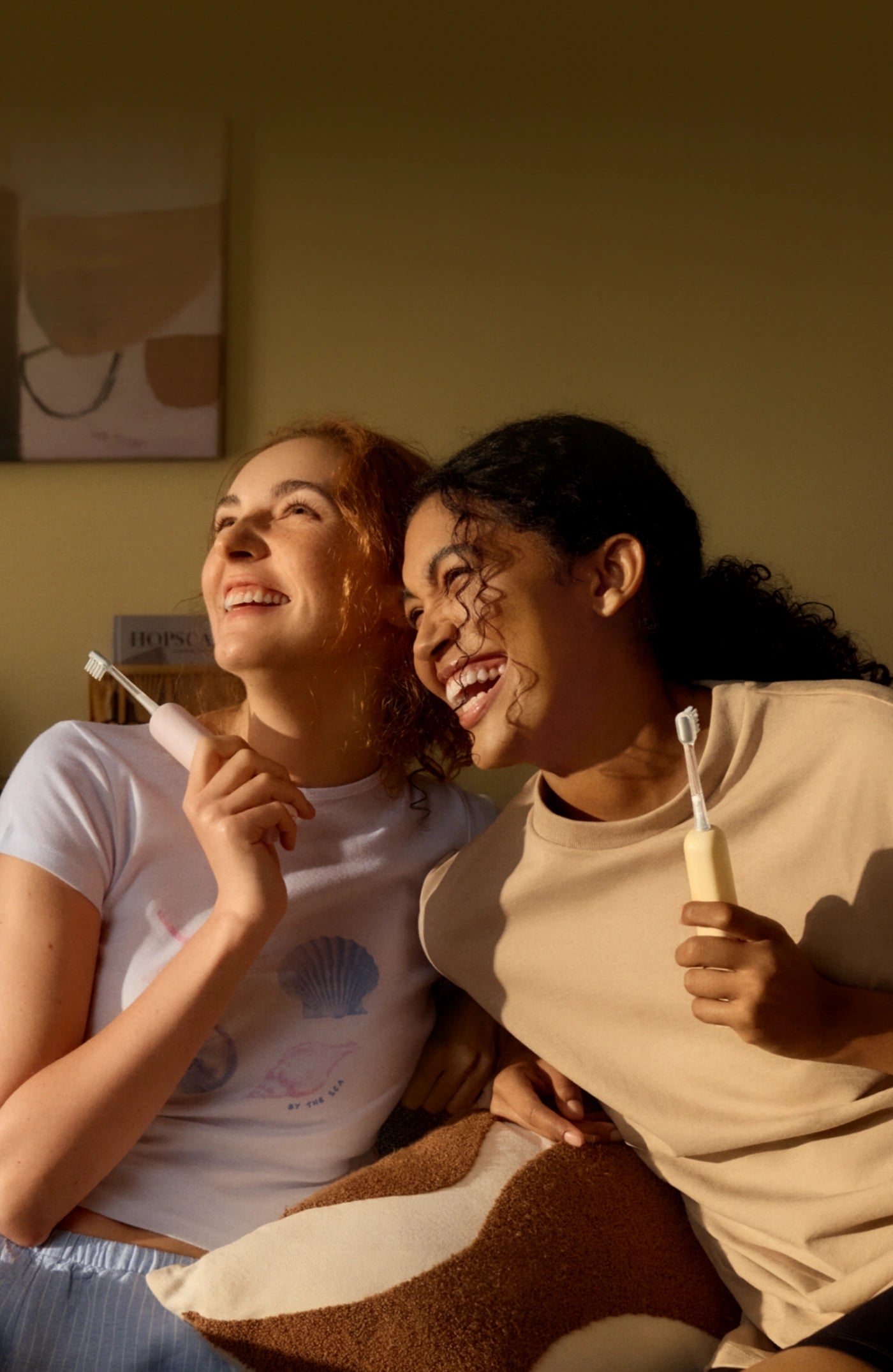 Two women sitting together, one holding a toothbrush and the other a toothpaste tube, in a warm indoor setting.